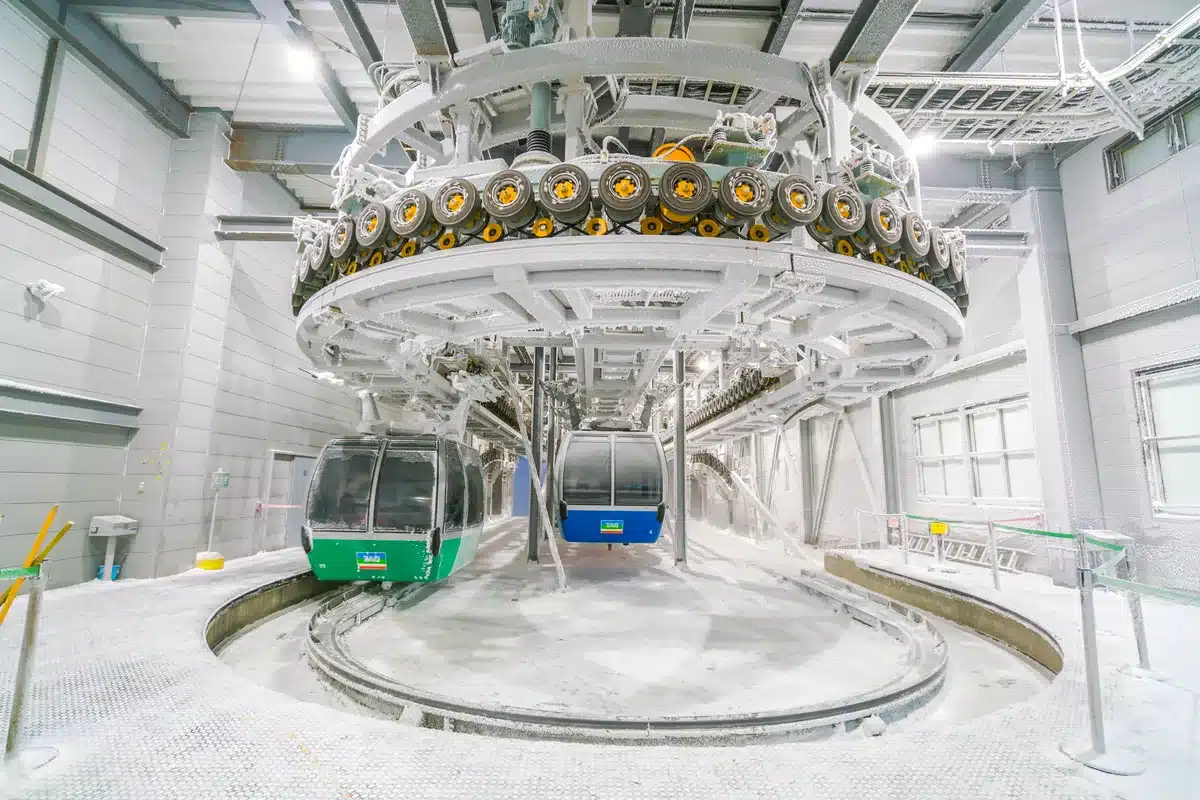 Estación de un teleférico con cabinas verdes y azules bajo la estructura de su rueda