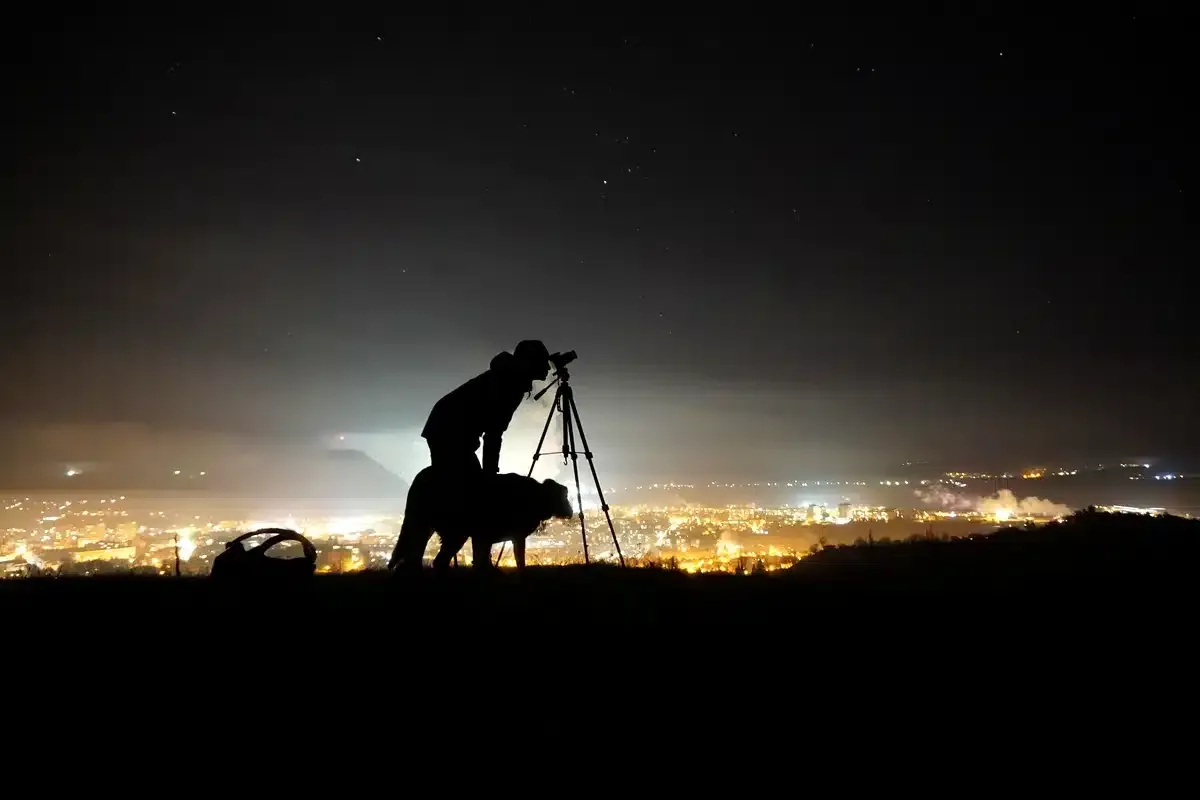 Silueta de una persona que observa con un telescopio desde una colina, con las luces de una ciudad al fondo