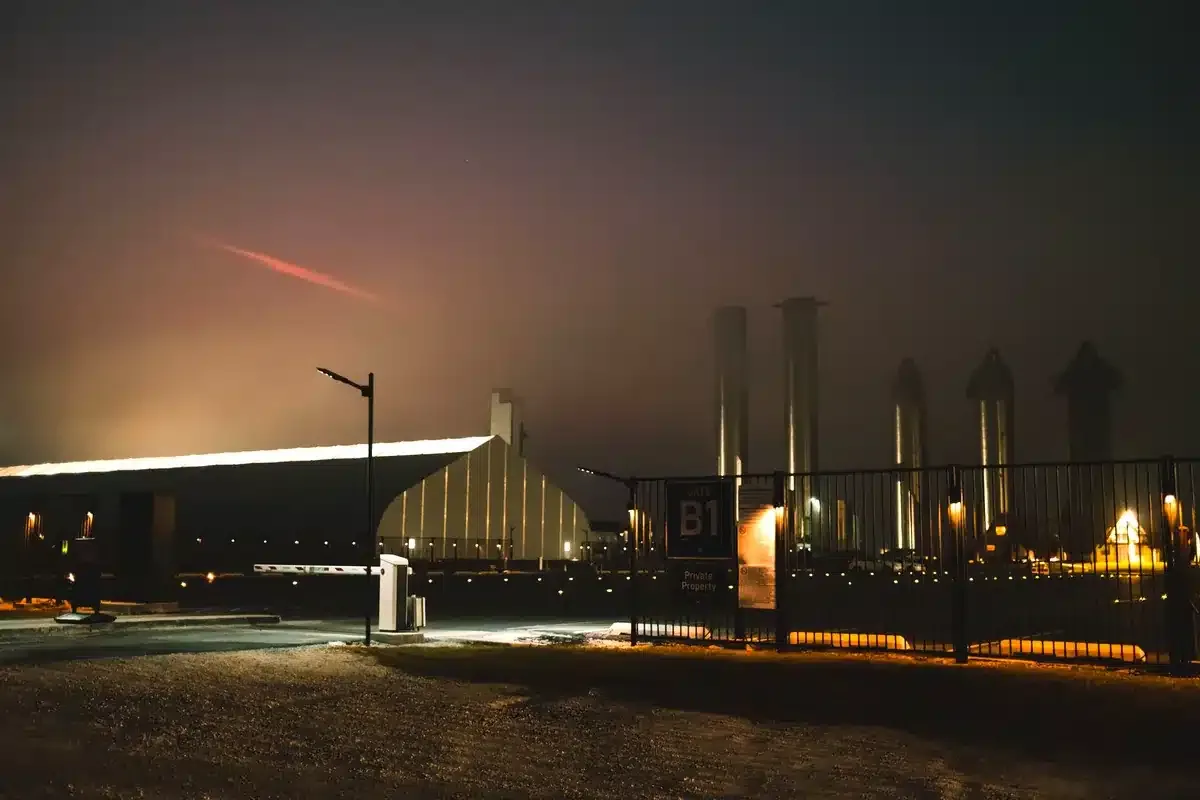 Base de lanzamiento de noche, con cohetes y tanques iluminados detrás de una cerca y un trazo rojo en el cielo