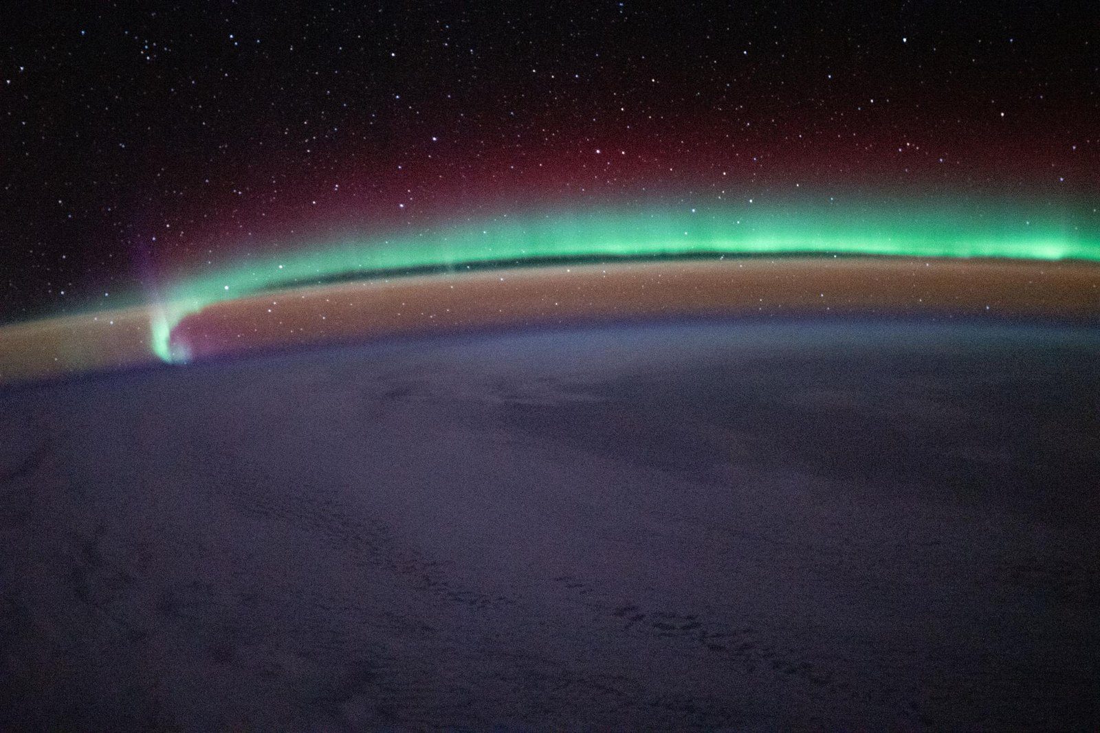 Aurora verde con un resplandor rojizo sobre el horizonte de la Tierra y un cielo lleno de estrellas, vistos desde la Estación Espacial