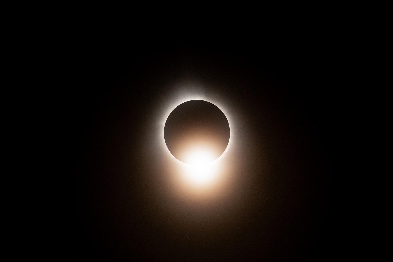 Eclipse total de Sol con la corona blanca alrededor de la Luna y un destello anaranjado en la parte inferior, efecto de anillo de diamante