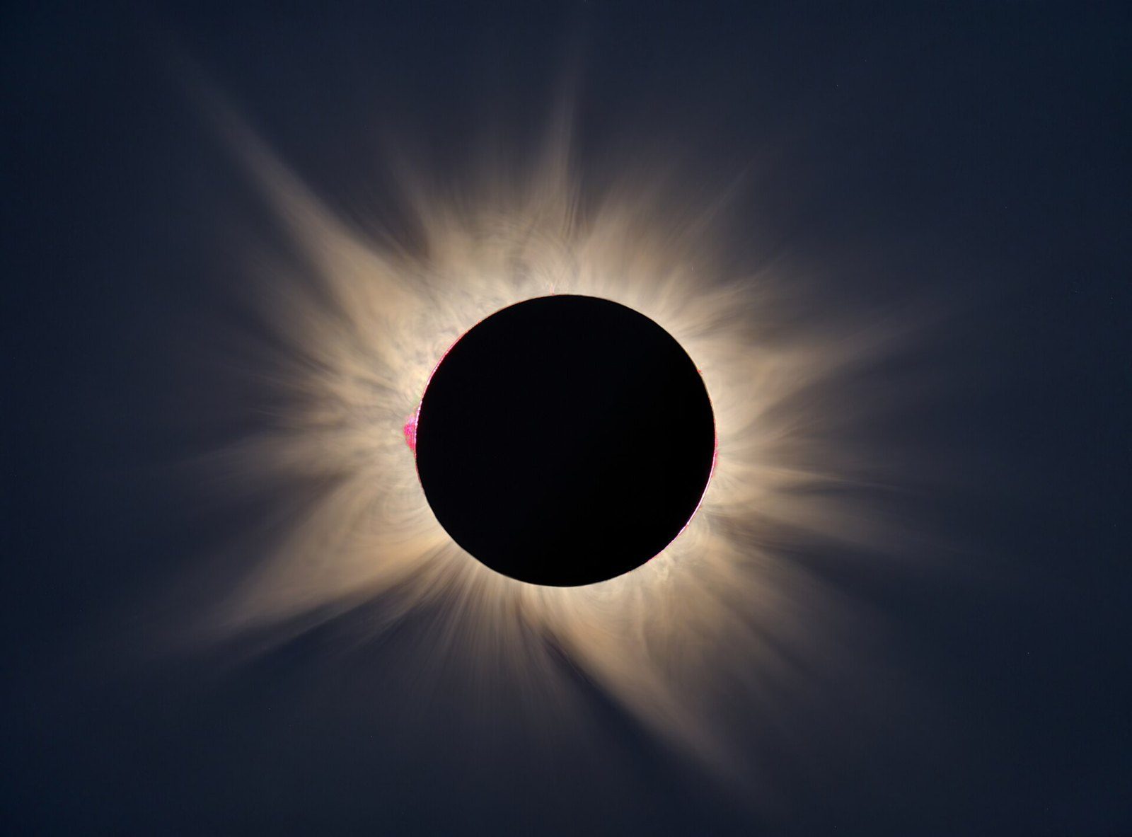 Eclipse total de Sol con la corona solar blanca y alargada rodeando el disco negro de la Luna sobre un cielo oscuro, con pequeñas protuberancias rosadas