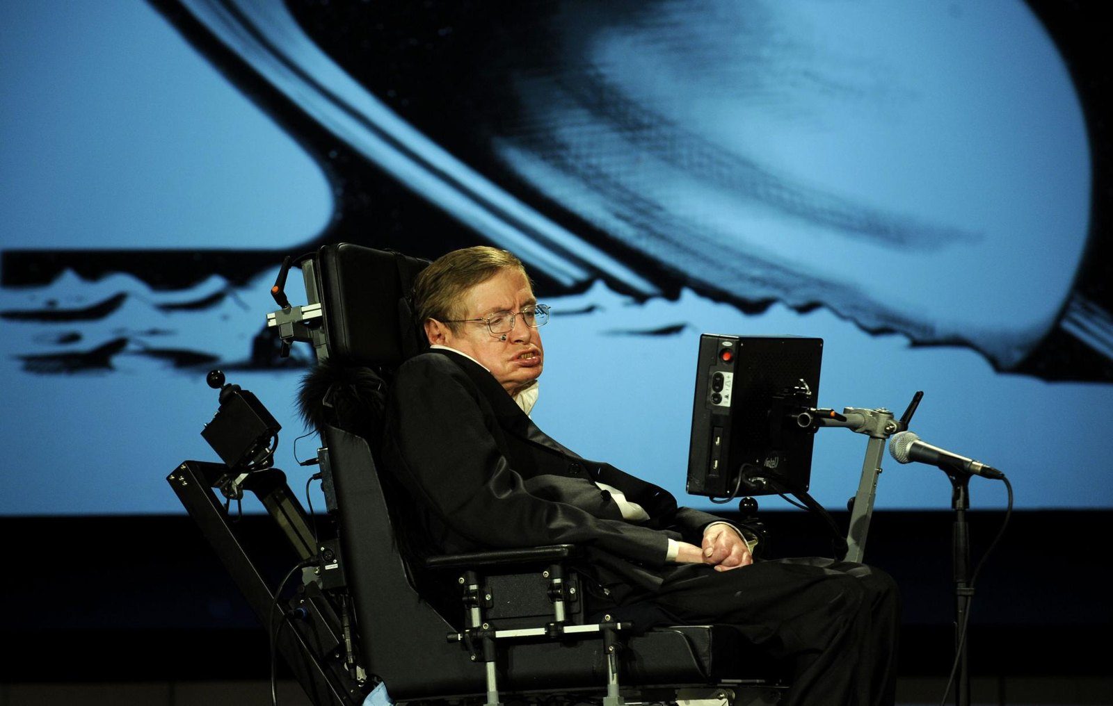 Stephen Hawking sentado en su silla de ruedas con un micrófono, frente a una pantalla azul con una imagen de Saturno y sus anillos